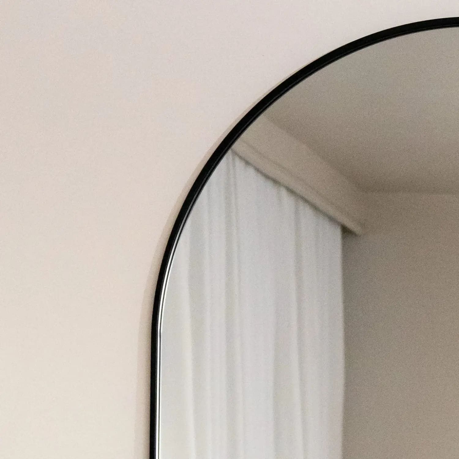 Arched mirror with a thin black frame reflecting white curtains.